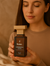Woman holding a bottle of 'Dastan Forbidden Aura' perfume against a warm-toned background